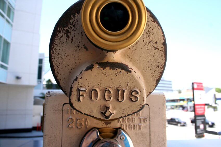 A close-up shows a coin-operated binocular viewer with "FOCUS" embossed on the front, set outdoors against a backdrop of buildings—ideal for Service Professionals like plumbers assessing new project sites.