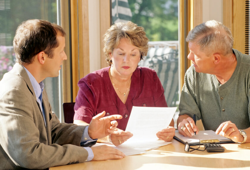 Three adults sit at a table and discuss their next project. One holds a document, another points to something on the table, and the third takes notes on a notepad. This scene is typical for service professionals like plumbers or electricians as they plan their work.