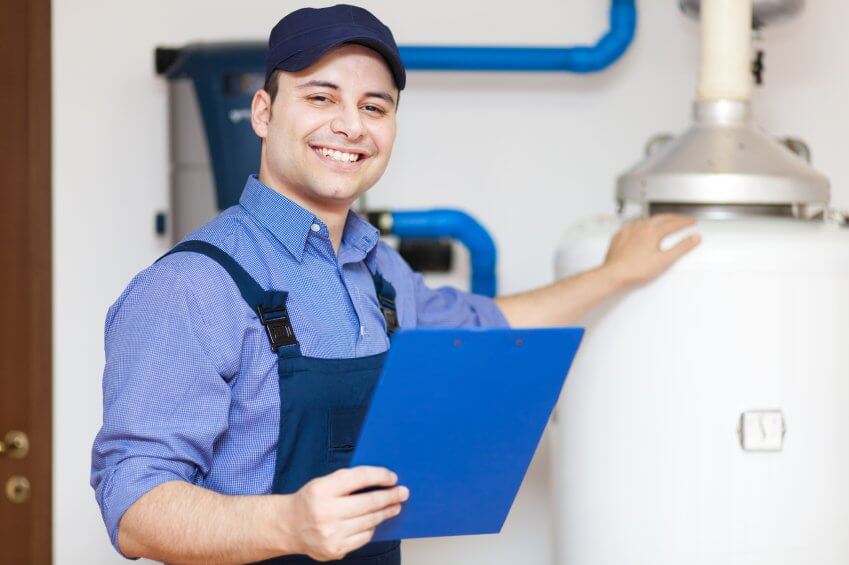 A smiling Service Professionals technician in uniform stands next to a water heater with exposed blue pipes and holds a blue clipboard.