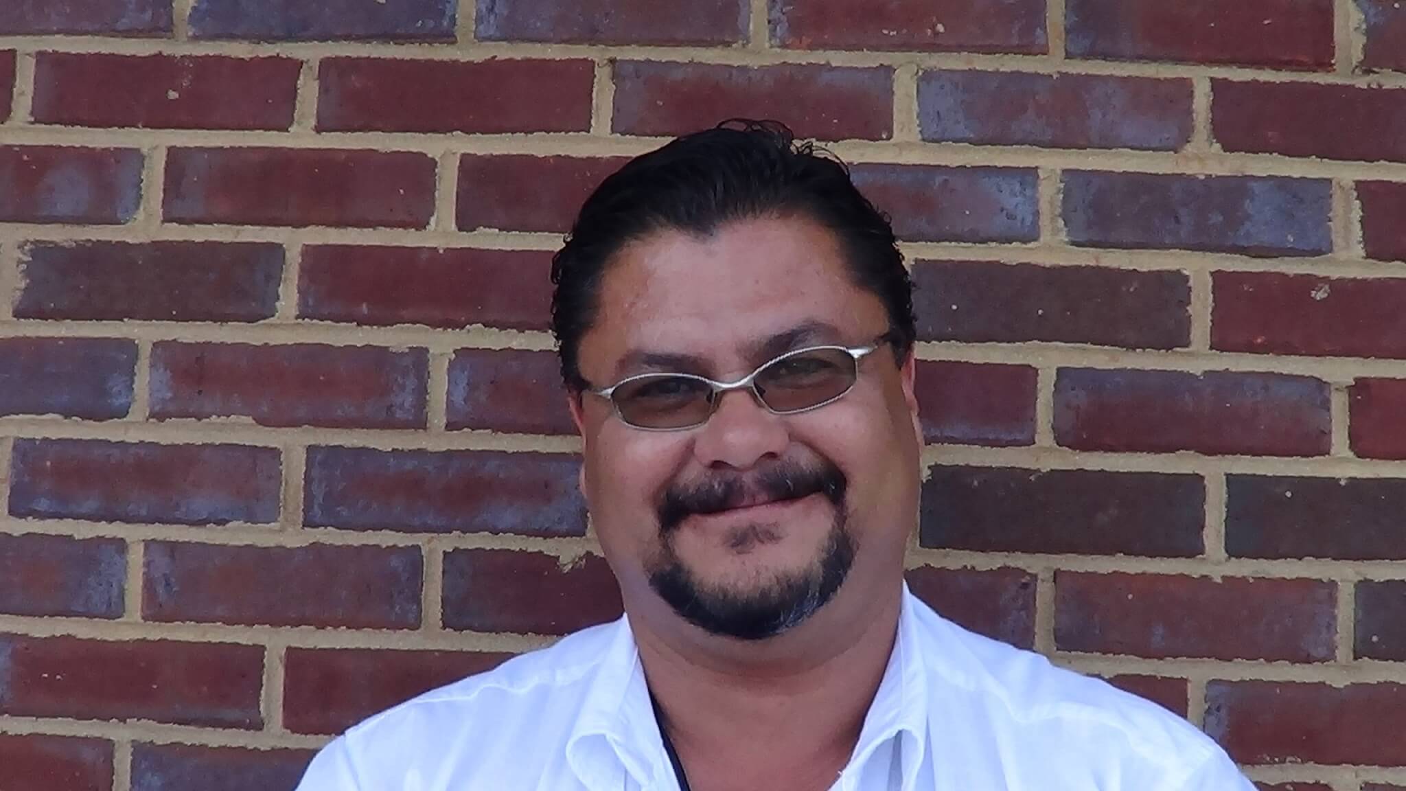 A man with dark hair, a goatee, and glasses wears a white shirt and smiles at the camera as he stands in front of a red brick wall. He looks like a skilled electrician ready for the job.