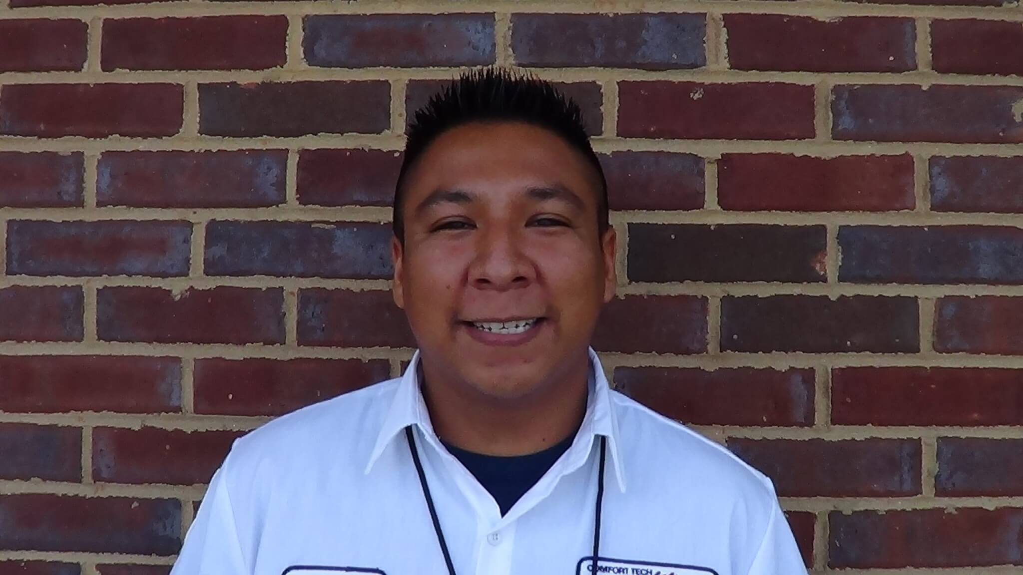 A man in a white collared work shirt stands in front of a brick wall, looks at the camera, and smiles—showcasing the friendly face of Service Professionals like electricians and plumbers.