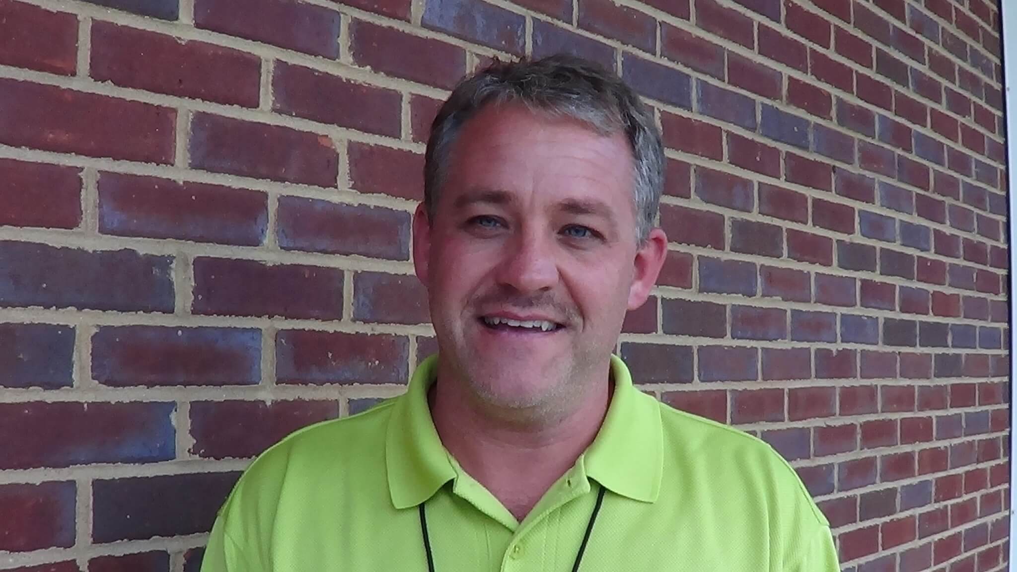 A man in a bright yellow polo shirt smiles in front of a red brick wall, ready to join local HVAC technicians and plumbers on their next job.