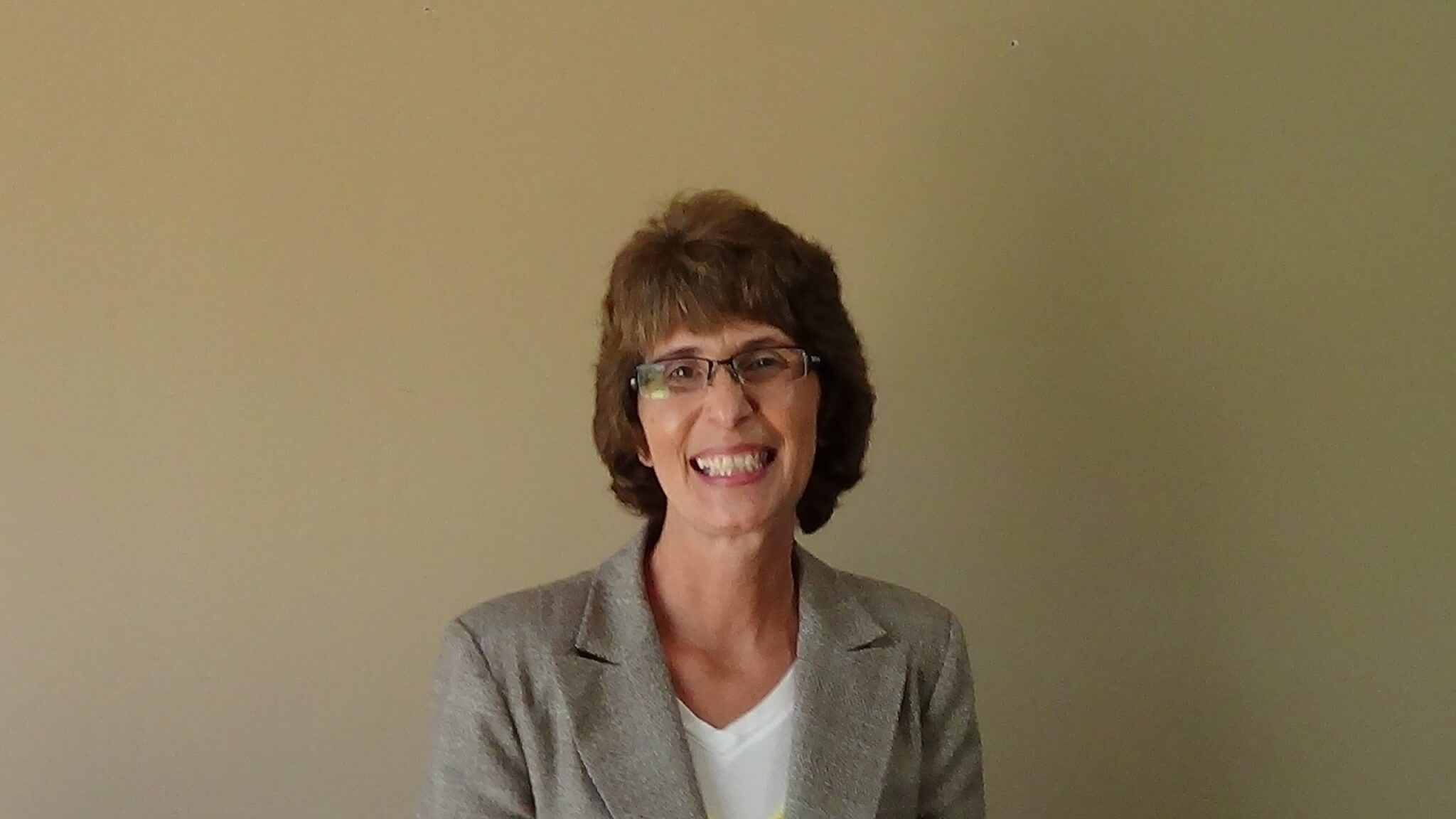 A woman with short brown hair and glasses, dressed in a gray blazer over a white top, smiles in front of a plain beige wall—an ideal look for service professionals such as electricians and plumbers.