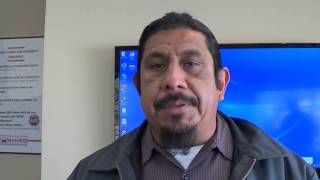 A dark-haired man with a goatee stands indoors in a collared shirt and jacket. He faces a blue computer screen and a wall sign, projecting the trustworthy look of a professional whom electricians or plumbers might rely on.