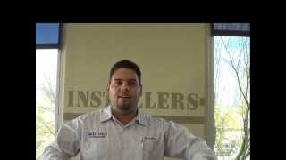 A man in a collared shirt stands indoors in front of a beige wall with the partial word "INSTALLERS" visible behind him. He represents skilled HVAC technicians.