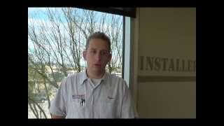 A man wearing a light gray work shirt stands indoors by a window overlooking trees. The wall beside him displays the partial word “INSTALLER,” suggesting he is likely a skilled service professional—possibly an electrician or plumber.
