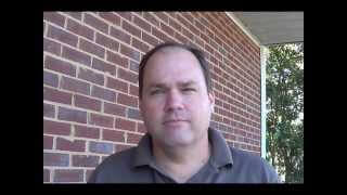 A man with short brown hair and a gray polo shirt stands outdoors in front of a red brick wall, looking like an HVAC technician.
