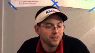 A person wearing glasses, a white visor cap, and a dark jacket—dressed like many HVAC technicians or plumbers—sits indoors in front of a wall covered with paper and blue tape.