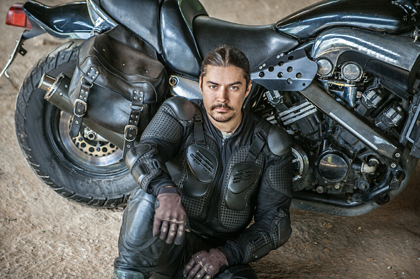 A man wearing black armored motorcycle gear sits on the ground next to a black motorcycle and looks up at the camera, appearing ready for action like top service professionals or on-call electricians.