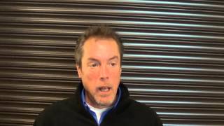 A middle-aged man with short hair and a trimmed beard, who looks like a skilled HVAC technician, stands in front of a metallic roller shutter and speaks.