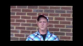 A man in a plaid shirt and baseball cap, resembling many skilled HVAC technicians, stands in front of a brick wall and looks at the camera.