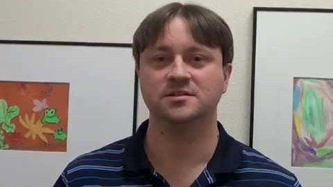 A man with short brown hair, wearing a navy striped polo shirt, stands in front of framed artwork on the wall. He looks like an electrician ready for work.