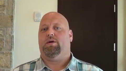 A bald man with a goatee and a plaid shirt stands indoors in front of a dark door and a light-colored wall. He looks like an experienced service professional—perhaps a plumber or an electrician.