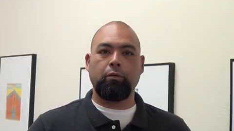 A man with a shaved head and dark beard, dressed in a black collared shirt, stands indoors like many HVAC technicians. He poses in front of framed artwork on a light-colored wall.
