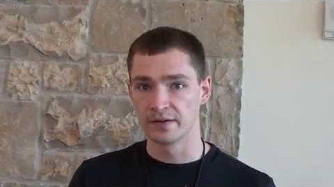 A man with short brown hair and a black shirt stands in front of a stone wall, looking confidently at the camera and embodying the presence of skilled Service Professionals.