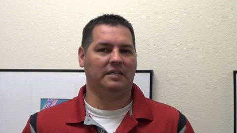 A skilled Service Professional with short dark hair and a red collared shirt stands in front of a light-colored wall decorated with framed pictures.