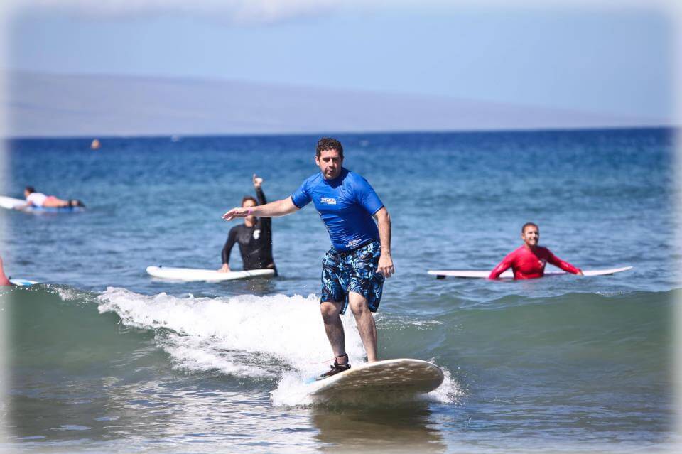 Wearing a blue shirt and patterned shorts, a skilled Service Professional surfs on a small wave as other surfers wait on their boards in the background.