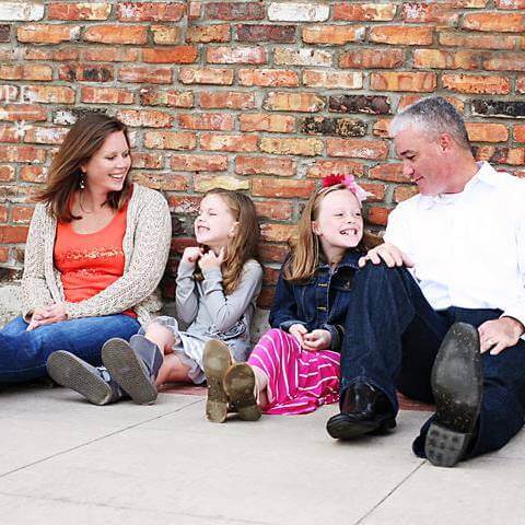 A woman, an electrician, and two young girls sit on the ground in front of a brick wall, smiling and talking together.