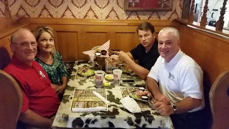Four adults—likely service professionals such as HVAC technicians or plumbers—sit together in a restaurant booth, sharing a meal. They smile and pose for the camera, with drinks and condiments on the table.