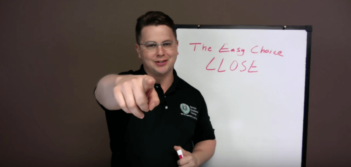 Wearing glasses and a black polo shirt, a person points at the camera in front of a whiteboard that reads "The Easy Choice CLOSE" in red marker. This tip helps HVAC technicians and plumbers improve their sales approach.