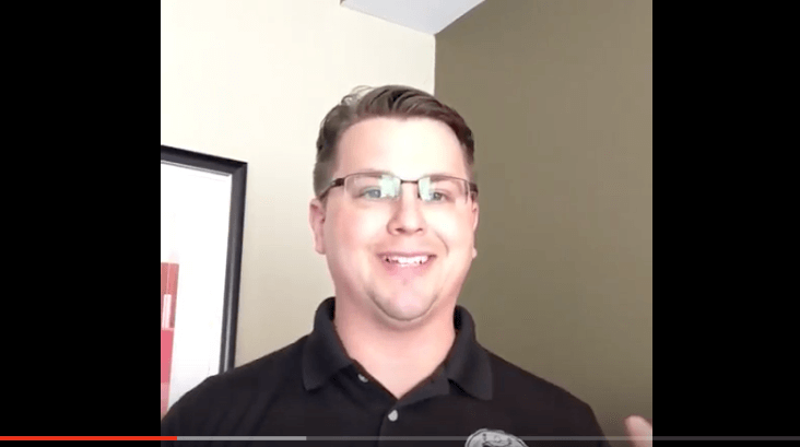 A man in glasses and a black polo shirt stands indoors, smiling at the camera. He looks like a skilled service professional—perhaps an HVAC technician or electrician. Behind him, a two-tone wall and a framed picture complete the scene.