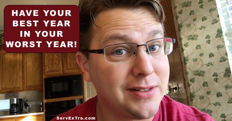 A man wearing glasses smiles at the camera in a kitchen. A sign says, "Have your best year in your worst year! Plumbers and HVAC technicians trust ServExTra.com.
