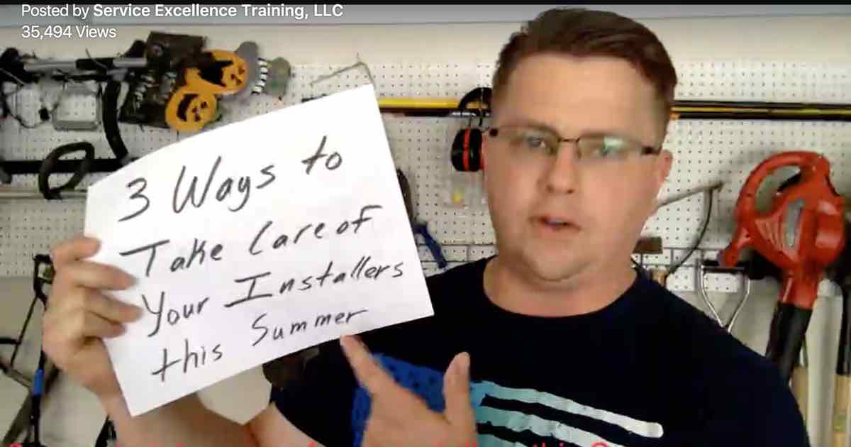 A man wearing glasses stands in a workshop with tools on the wall, holding a handwritten sign that reads, "3 Ways to Take Care of Your Installers this Summer." He offers tips especially useful for HVAC technicians and other service professionals.