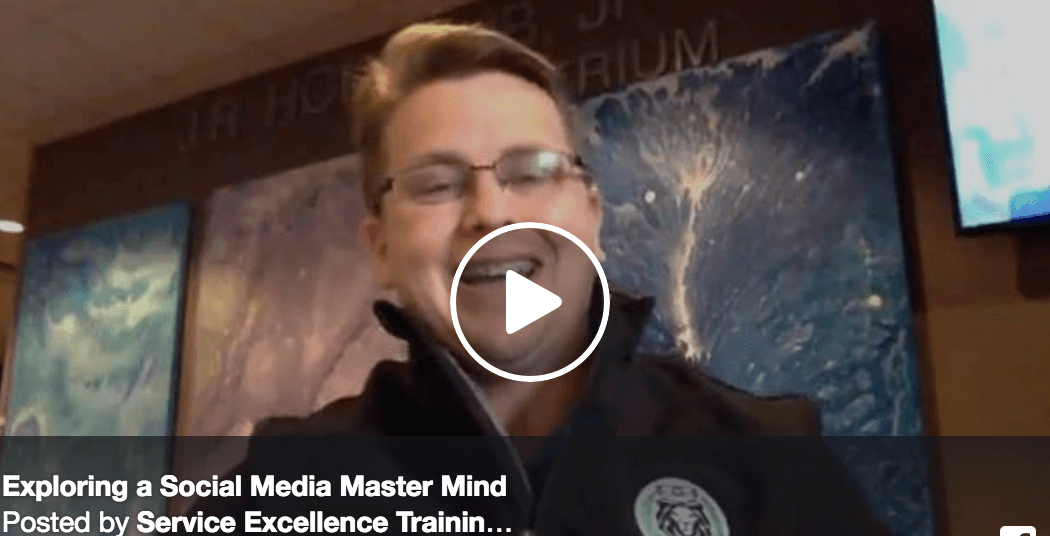 A person in glasses and a dark jacket speaks in front of abstract blue artwork. The video, titled "Exploring a Social Media Master Mind," highlights coaching and service excellence in the digital era.
