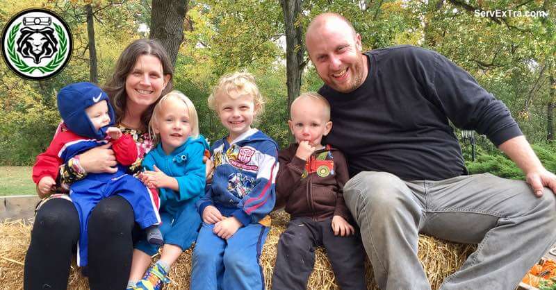 A woman and a man sit with four young children on straw bales outdoors. They all smile warmly at the camera, radiating Service Excellence. Trees and lush greenery create a beautiful backdrop.