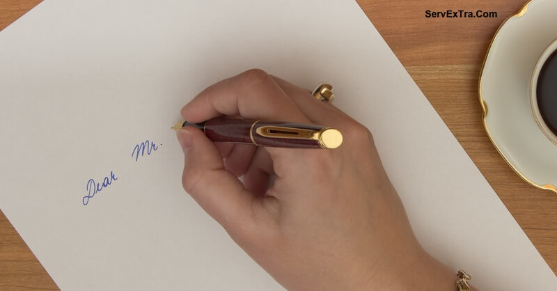 A hand grips a fountain pen and writes "Dear Mr." in blue ink on white paper. A cup of coffee sits nearby on the wooden table, creating a personal, inviting setting for home services or coaching correspondence.