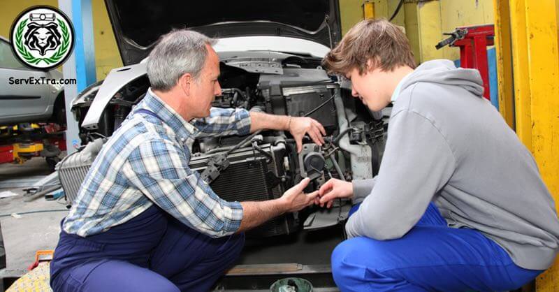 An older mechanic and a younger mechanic work together on a car engine in a workshop, demonstrating Service Excellence. Tools and car parts surround them.