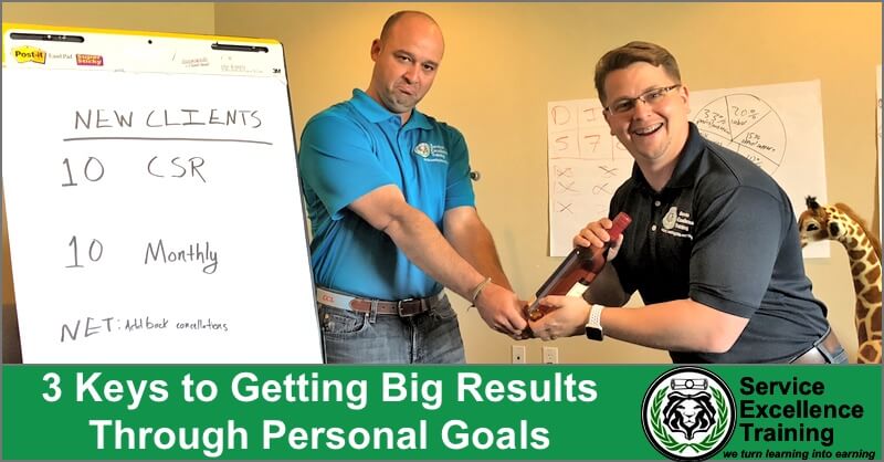 Two men stand in a classroom, holding a baton together during a service training session. One stands next to a flip chart that lists new client goals. A giraffe toy, the training logo, and the seminar title appear in view.