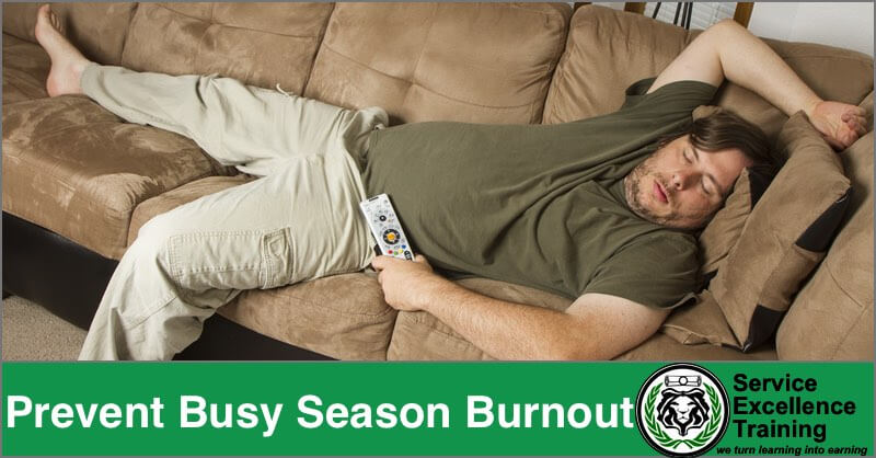 A man sleeps on a couch, holding a remote control. The text says, “Prevent Busy Season Burnout.” The Service Excellence Training logo appears, promoting coaching for home services professionals.
