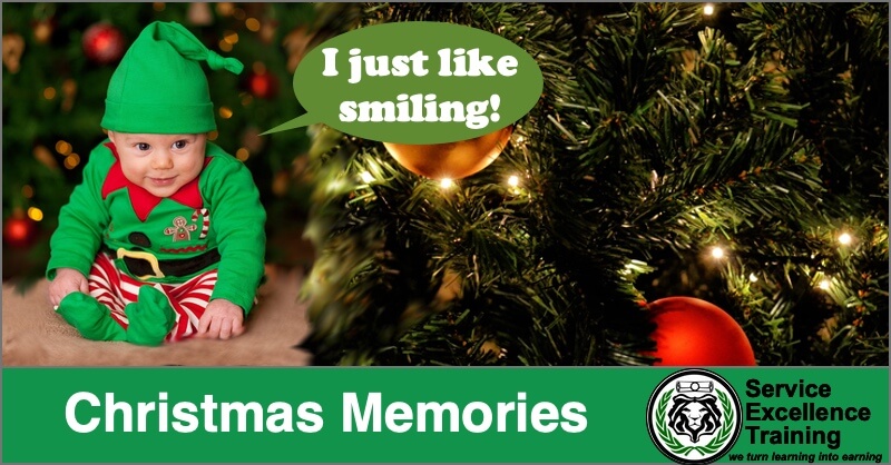 A baby in an elf costume sits by a Christmas tree. A speech bubble says, "I just like smiling!" The text below reads: "Christmas Memories." The Service Excellence Training logo for Service Professionals and electricians appears underneath.