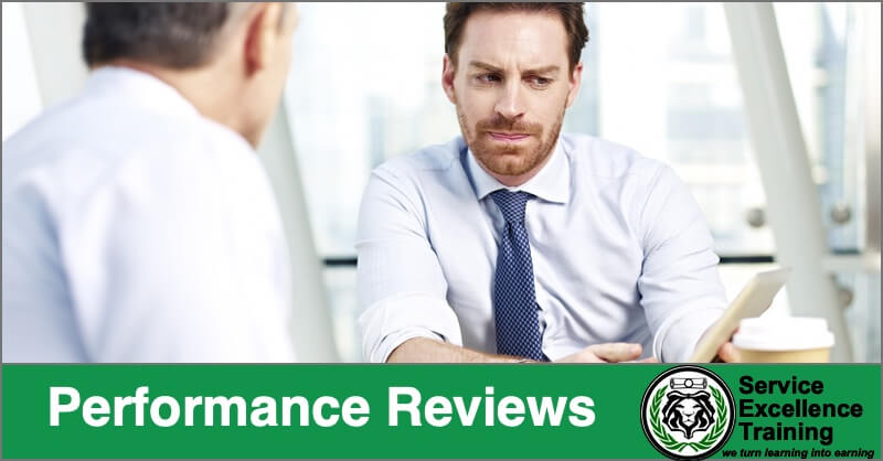 Two men in business attire sit across from each other at a table, having a serious discussion. A green banner labeled "Performance Reviews" with the Service Excellence Training logo highlights growth opportunities for service professionals such as electricians and HVAC technicians.