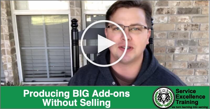 A man wearing glasses stands outside by a brick wall and says, “Produce BIG Add-ons Without Selling—Service Excellence Training for plumbers and electricians.” A play button appears at the center of the image.