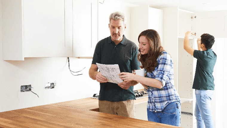 Three people work in a kitchen. Two review papers, and an electrician installs cabinets in the background.