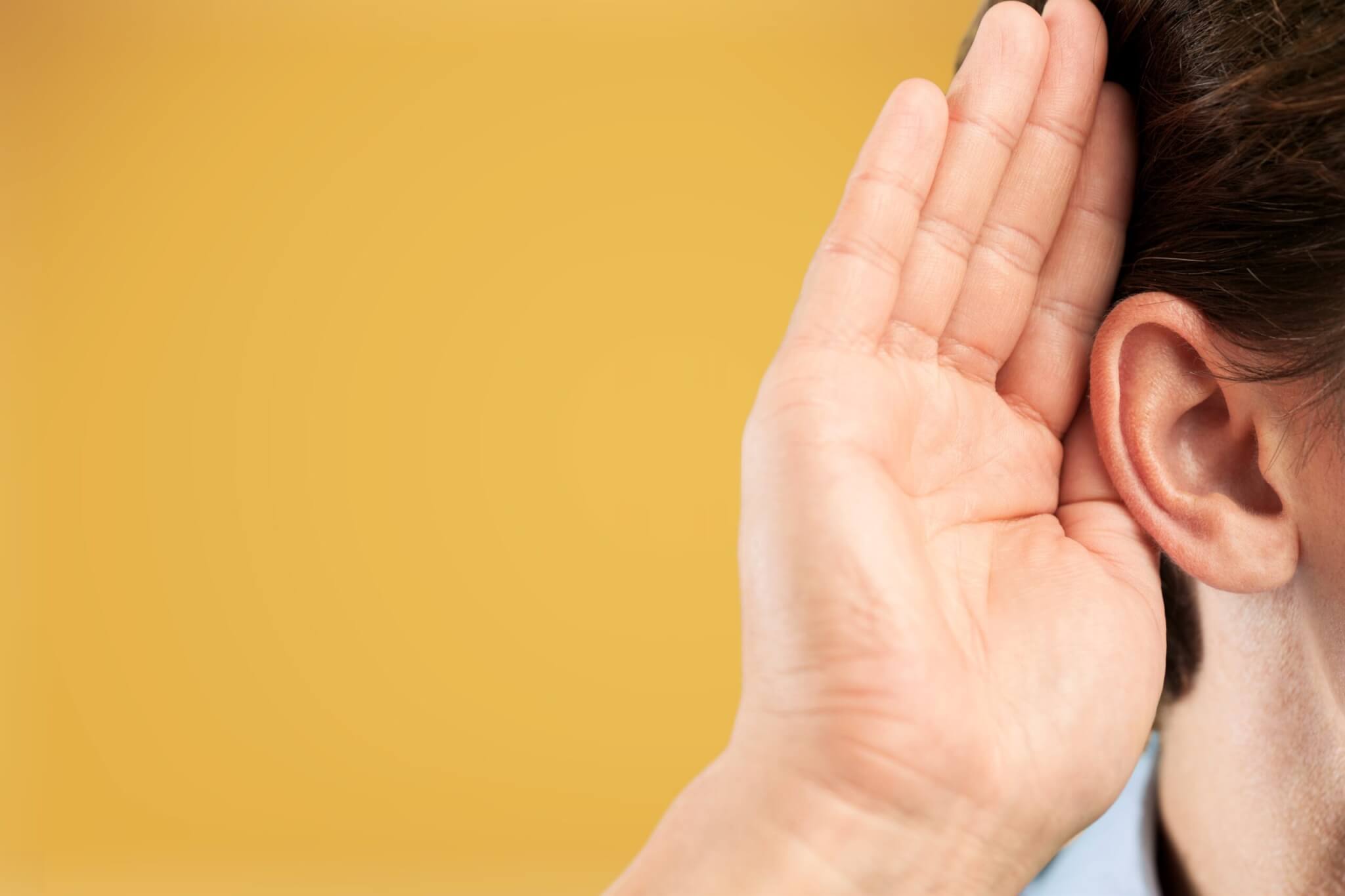 A person holds their hand to their ear, as if listening closely, to symbolize the importance of Service Excellence, against a plain yellow background.