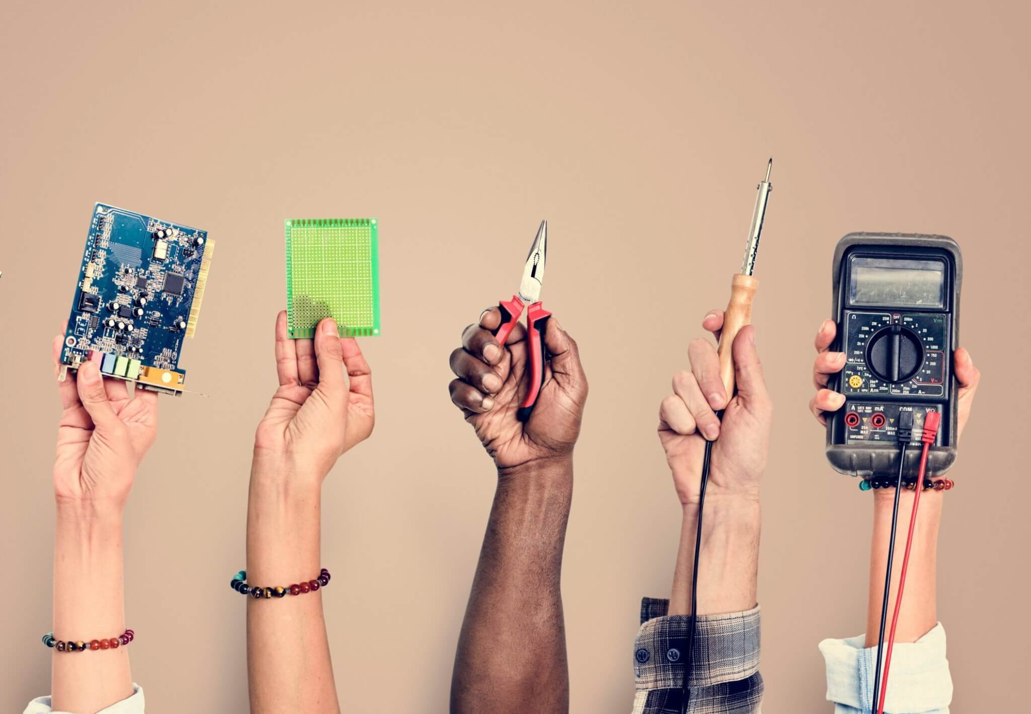 Four people hold up a circuit board, a prototyping board, pliers, a soldering iron, and a digital multimeter—essential tools for service training and achieving Service Excellence in the home services industry.