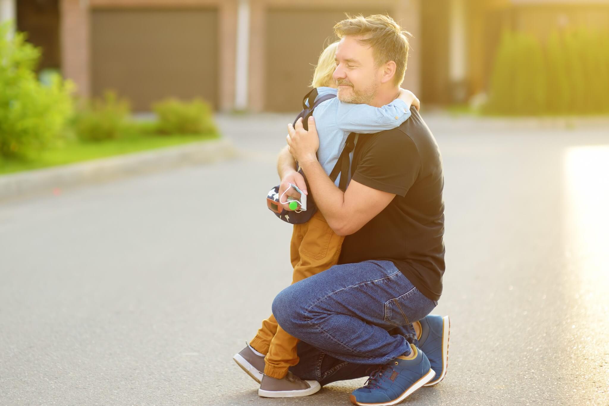 A home service business owner kneels on a street and hugs a small child holding a toy car. Surrounded by greenery and garages, they share a warm family moment that goes beyond work or coaching.