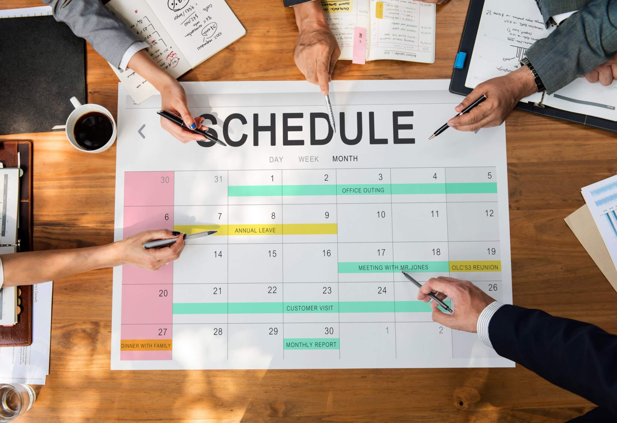 Four people sit around a wooden table, pointing to a large printed calendar labeled "Schedule" as they plan service training sessions for home service business owners. Notes, documents, and a cup of coffee lie on the table.