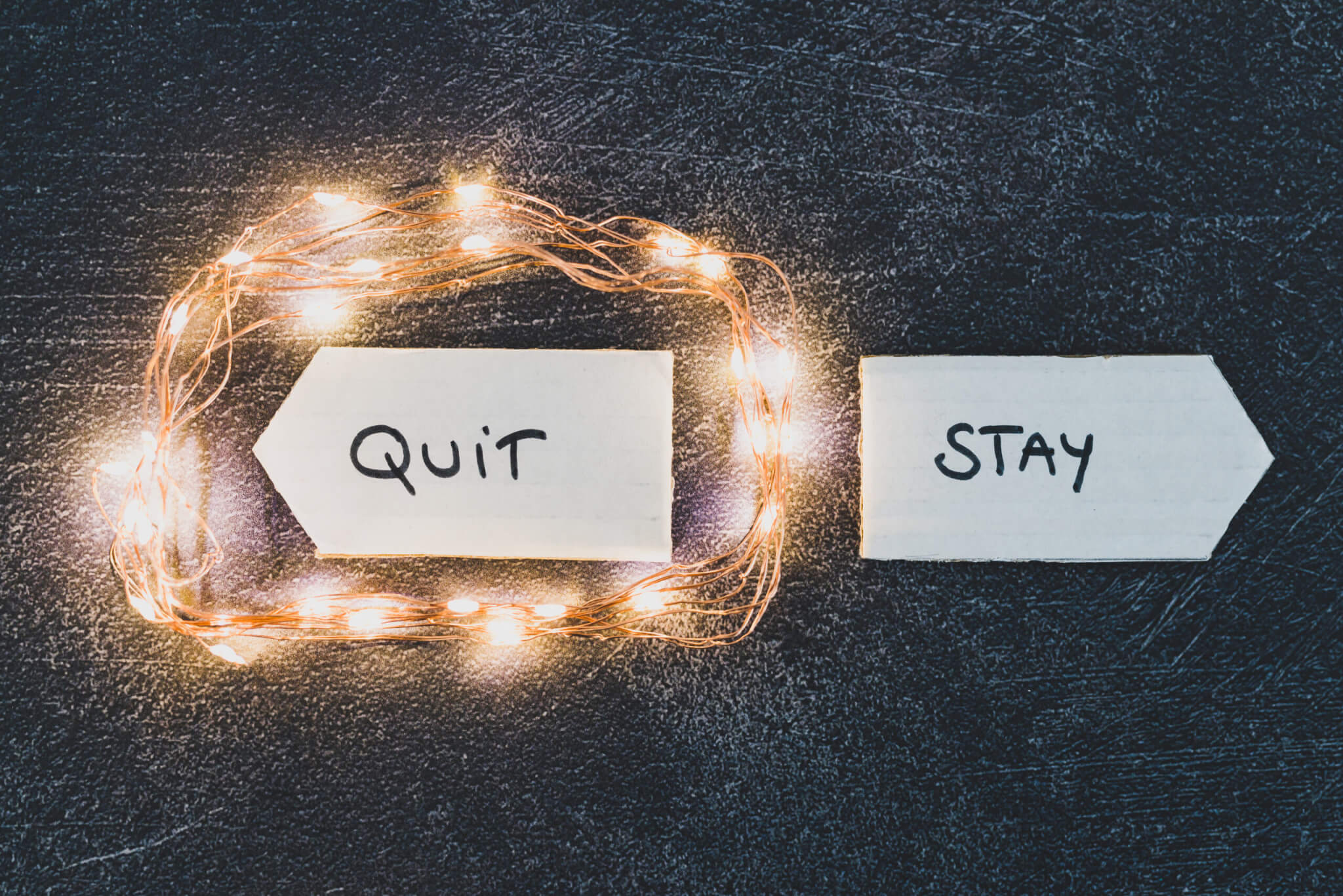Two arrows sit on a dark surface. One, labeled "QUIT," glows with string lights. The other, labeled "STAY," remains plain. This stark image captures the choice home service business owners face as they decide their next move in the industry.