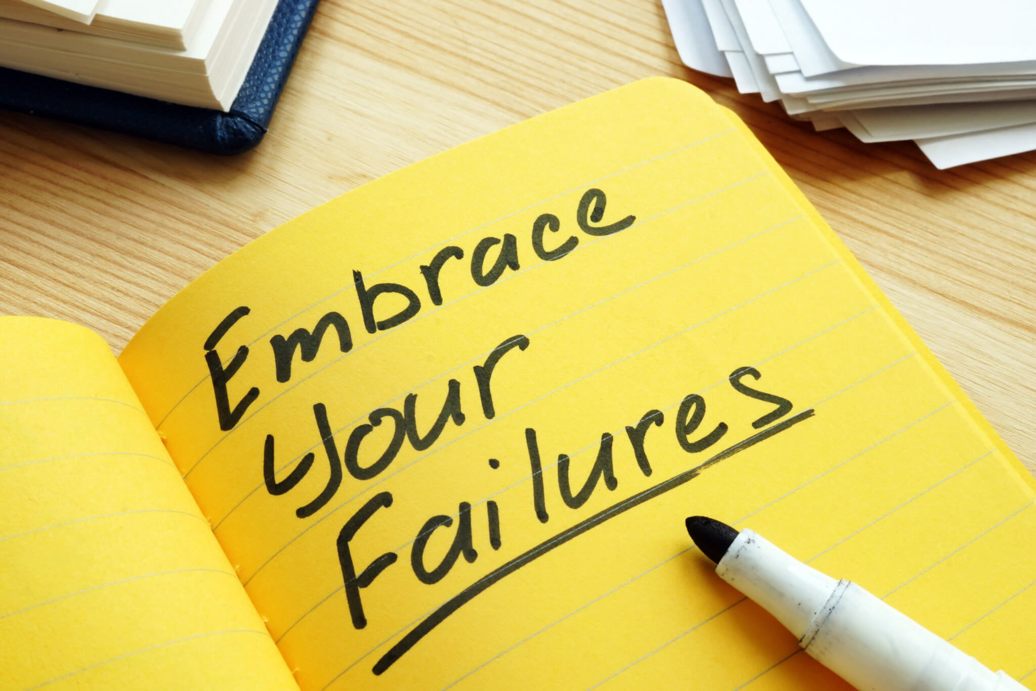 A yellow notebook displays the handwritten message "Embrace Your Failures," with an uncapped marker resting on the page. Stacks of paper sit in the background, making this scene a perfect source of inspiration for home service business owners or anyone seeking coaching.