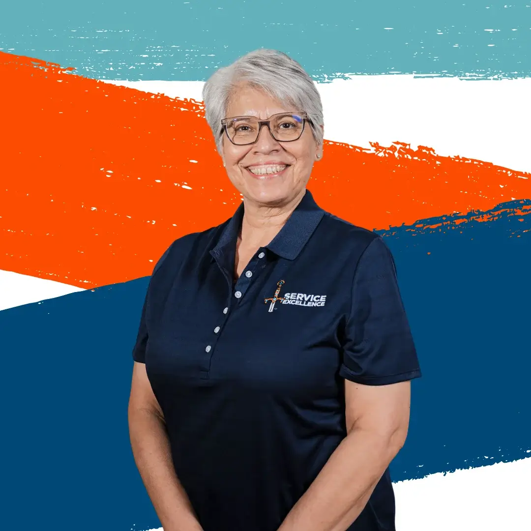 A smiling older woman with short gray hair and glasses wears a navy blue "Service Excellence" polo as she stands before a colorful abstract background, showcasing her passion for service training and coaching in home services.