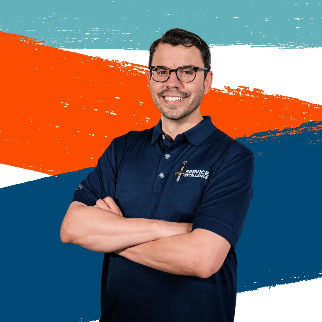 A man with glasses smiles confidently, arms crossed, wearing a navy polo shirt with a "Service Excellence" logo. He stands in front of a colorful abstract background, proudly representing service training in the home services industry.