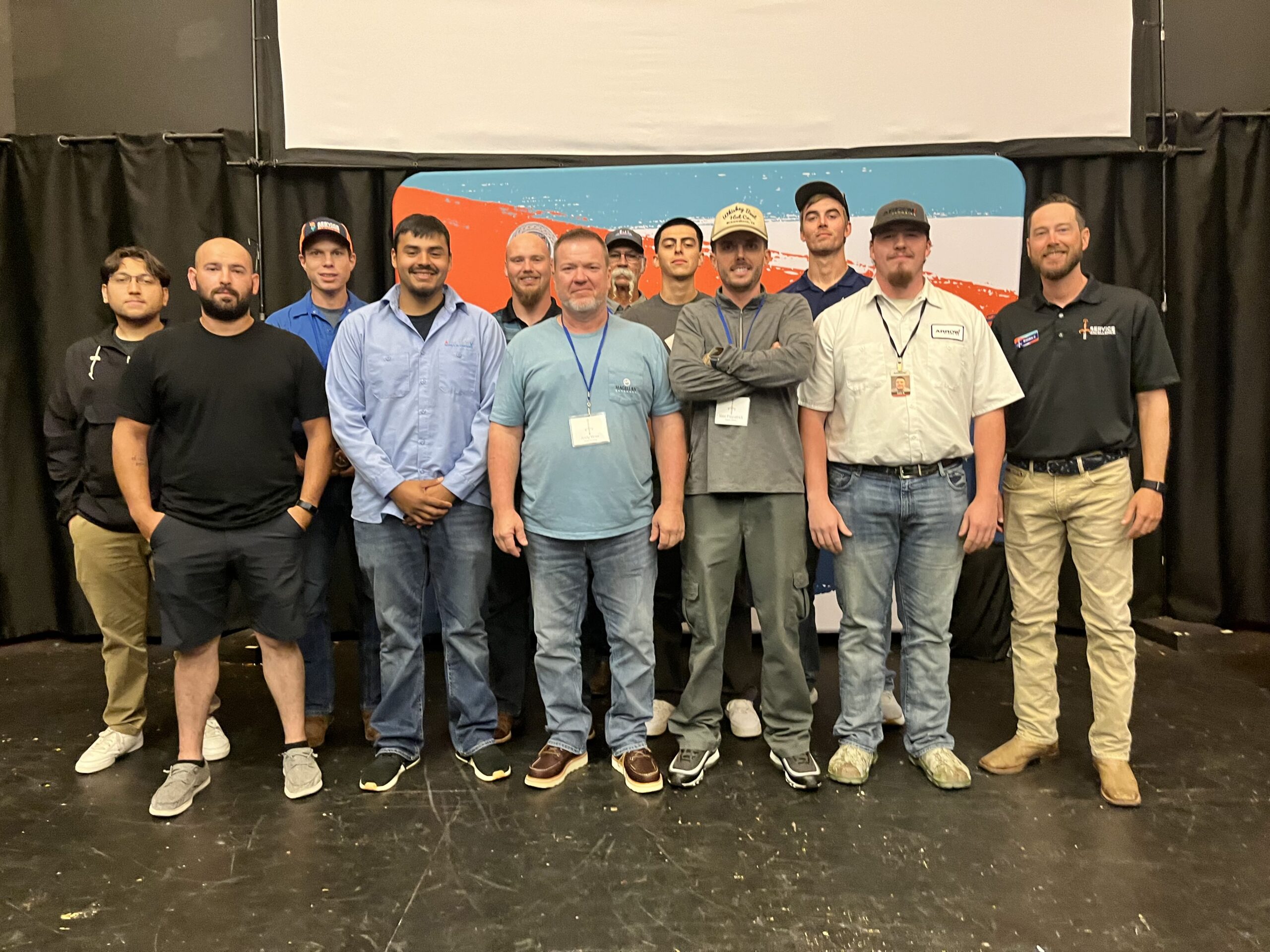 Twelve men stand together indoors in front of a colorful backdrop and pose for a PRESS PLAY Training Series group photo.